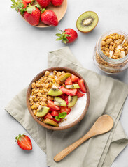 Natural yogurt with granola, kiwi and strawberries in a wooden bowl on a light background with fresh berries, fruits and napkin.