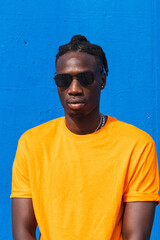 Serious black man with sunglasses and trendy orange t-shirt looking at camera against blue background