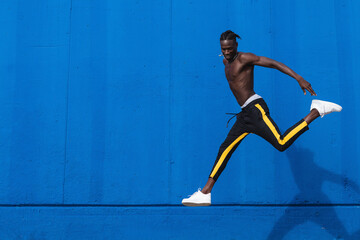 Full body side view of happy young shirtless African American sportsman running and jumping near bright blue wall