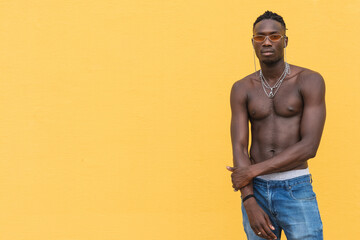 Serious young shirtless African American male with metal necklaces posing against yellow wall