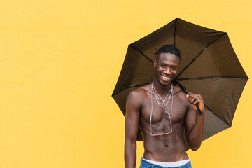 Optimistic black man shirtless smiling happily while holding an umbrella against yellow wall enjoying sunny summer day