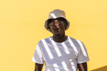 Modern hipster African American guy with striped shadow on white t shirt wearing sunglasses and panama hat and looking at camera while standing against yellow wall