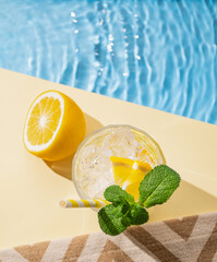 Cold lemonade in a glass with lemon, mint and ice on a yellow background with the edge of a pool...