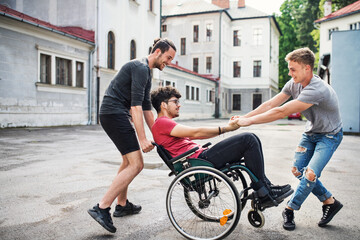 Young man in wheelchair spending free time outdoor with friends, having fun in city. Male...