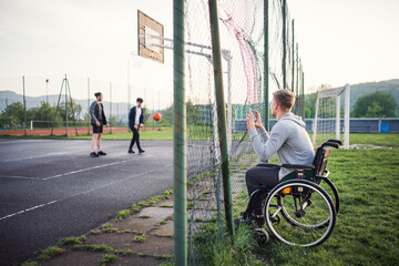 Fototapeta premium Disabled young man feeling excluded, want to play basketball with his friends, but because of wheelchair he can't.