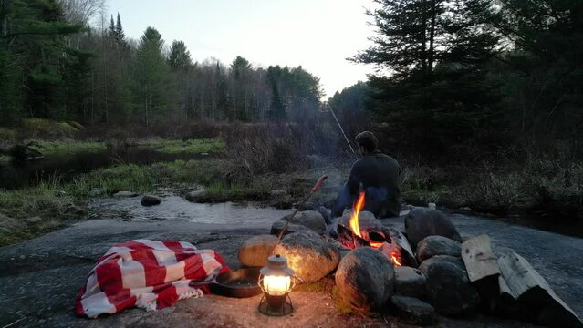 Man fishing by a campfire with a lantern and blanket in a forest at twilight, surrounded by trees and the tranquil sounds of nature.