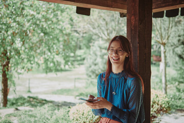 Joyful teenage girl using smartphone in a rustic wooden gazebo, surrounded by a lush garden....