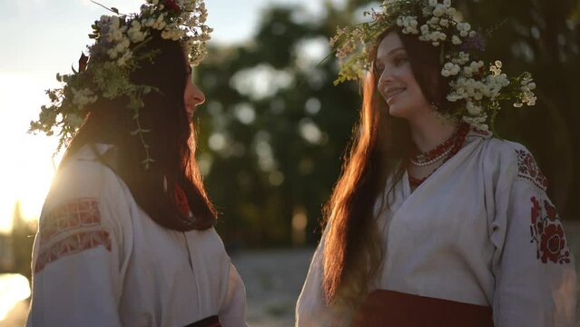 Slow motion. On the night of Ivan Kupala, two beautiful Ukrainian women in traditional embroidered shirts with wreaths of wild herbs and flowers look at each other and smile, standing outdoors on the