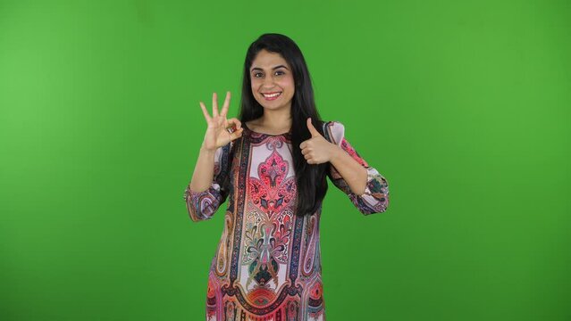 A portrait of an Indian girl making an okay sign from fingers on the green screen - green screen concept  sign language. An attractive girl making thumbs-up signs from one hand - hand movements  ge...