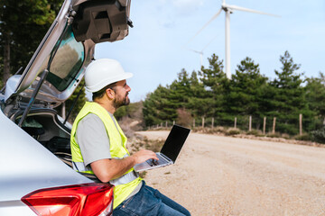 A wind turbine technician in a reflective vest and hard hat works on a laptop while sitting on a...