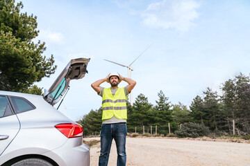 A wind turbine technician in a high-visibility vest and hard hat stands next to his car, preparing for work, with wind turbines in the nearby background. © Koldo_Studio
