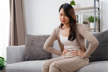 The woman with a stomachache is holding her stomach in pain, sitting on the sofa in the living room.