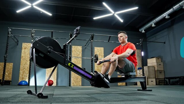 Man rowing on rowing machine in gym. Functional training. Sportsman doing cross training indoors. Man wearing red shirt and shorts. Gym is well lit and has modern design. 4K, UHD
