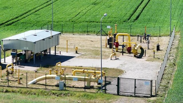 In the middle of a crop field, a gas station with yellow pipes, valves, and utility buildings. The fenced area houses an underground gas pipeline.