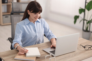 Woman taking notes during webinar at wooden table indoors