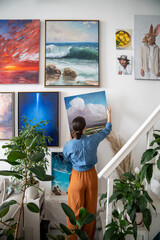 Interested woman painter hang newly painted modern canvas painting on wall in spacious art workshop with live houseplants. Actual solutions by decoration in contemporary interior design. Home gallery.