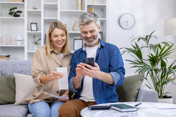 Happy couple managing their finances at home, using a smartphone to review expenses and receipts...