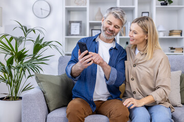 Obraz premium Smiling middle-aged couple sitting on a couch, enjoying a moment together while looking at a smartphone. Modern living room background.
