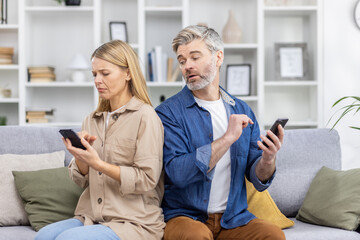 Couple sitting on couch looking upset while using smartphones, highlighting relationship issues and technology's impact on communication.