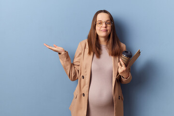 Puzzled confused pregnant woman wearing dress and jacket isolated over blue background holding...