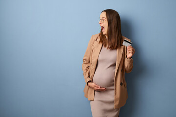Surprised pregnant woman wearing dress and jacket isolated over blue background looking away at...