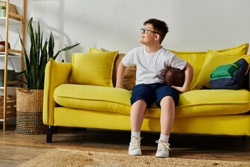 A charming boy with Down syndrome holding a football while seated on a yellow sofa.