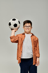 A little boy with Down syndrome holds a soccer ball on plain backdrop.