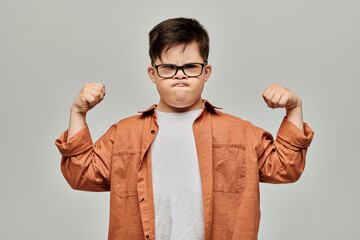 A little boy with Down syndrome with glasses flexes his muscles, showcasing his strength and determination. © Bliss