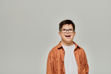A little boy, with Down syndrome, wearing glasses, stands against a gray background. © Bliss