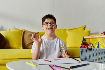 A adorable boy with Down syndrome in glasses happily drawing on a yellow couch.