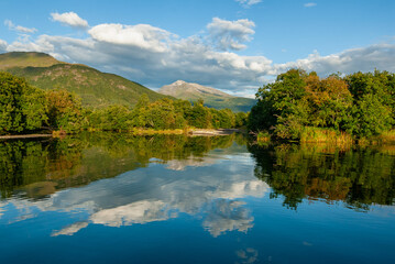 Tranquil lake surrounded by lush greenery with mountains in the backdrop, under a clear blue sky with fluffy white clouds reflected on the waters surface.