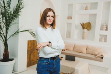 A pretty woman with glasses is standing in the middle of a bright living room, wearing a white t-shirt and jeans. Young confident woman working remotely.
