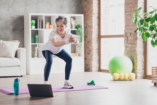 Full length photo of nice pensioner woman squats online training sportswear home exercising routine living room healthy lifestyle