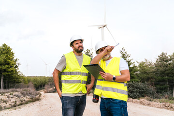Two wind technicians in reflective vests and helmets discuss a project with a clipboard, near...