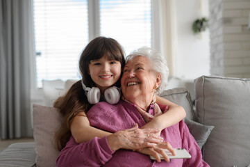 Cute girl hugging gradmother from behind, looking at camera, smiling. Portrait of an elderly woman spending time with granddaughter.