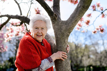 Portrait of elderly woman standing by magnolia tree, in park, having relaxing moment. Grandmother enjoying warm spring weather.