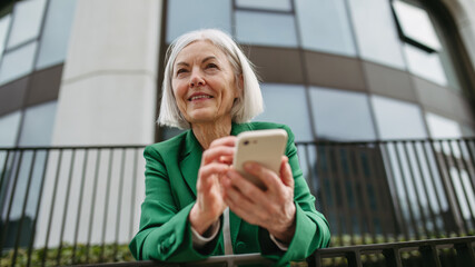 Mature businesswoman holding smartphone, waiting for business partner in the city. Beautiful older...