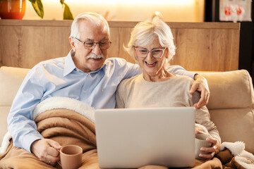 Smiling senior couple is relaxing at cozy home with mugs of tea in hands and movie a laptop.