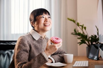 A happy asian freelancer sitting at home office, eating doughnut and smiling at computer monitor.