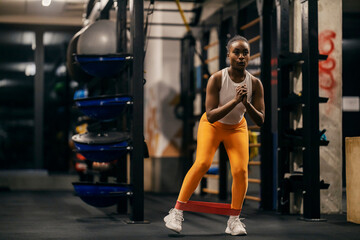 A black muscular black sportswoman is practicing her legs with resistance band at gym.