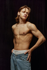 Naklejka premium Handsome young man standing shirtless, with muscular, relief strong body posing in jeans against black background. Concept of beauty, body, sport and health, fashion, wellness