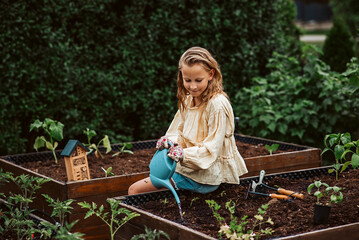 Girl watering small vegetable plants in raised bed, holding plastic watering can. Taking care of garden and planting spring flowers.