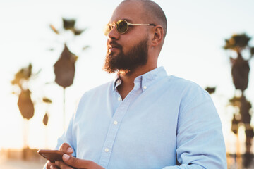 Handsome bearded male holding smartphone using 4G connection in roaming for chatting, hipster guy...