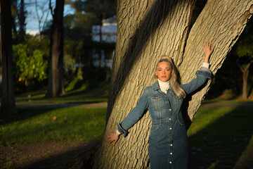Woman, young, pretty, blonde, with blue eyes and denim dress, leaning on the trunk of a large tree...