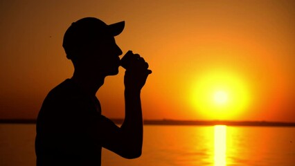 A man stands tall, silhouetted against a vibrant orange sunset, sipping his coffee and taking in the breathtaking view. The vastness of the sky and water creates a sense of awe and wonder.
