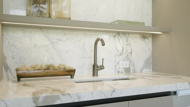 Elegant kitchen countertop with marble finish. Stainless steel sink with high curved faucet. Above countertop under-cabinet range hood. Tiled wall matching cabinets