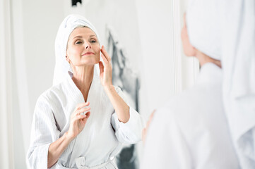 Mature woman looking at mirror and putting on face skin nourishing serum.