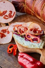 Sliced sausage with dried tomatoes lies on a wooden table. In the foreground is a sandwich made of this sausage and vegetables. Meat production.