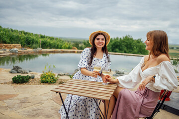 Two young smiling women are sitting at table and drinking champagne near pond.