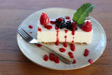 Cheesecake slice decorated with pieces of red and black berries, a mint leaf, and raspberry sauce. The dessert is served on a white plate with fork, putted on a wooden table.